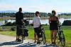 Tre personer med sykler stopper ved en landevei og ser utover en innsjø og grønne jorder.