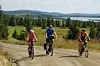 Tre personer sykler på en grusvei i grønt fjellandskap med innsjø bak.