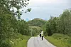 Syklister på landevei omgitt av grønn skog og åser en overskyet dag