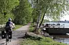 Syklist på grusvei ved elv med grønne trær og brygge med båt