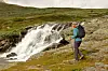 Vandrer med ryggsekk går forbi et lite fossefall i fjellet