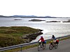 To personer sykler langs en kystvei med fjord og små øyer i bakgrunnen.