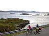 To personer sykler langs en kystvei med fjord og små øyer i bakgrunnen.