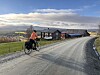 Person på sykkel langs grusvei ved gamle gårdsbygninger i åpent landskap