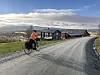 Person på sykkel langs grusvei ved gamle gårdsbygninger i åpent landskap