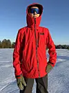 Person i rød Helly Hansen-jakke står på åpent snøområde i solskinn.