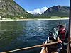 To personer i en liten båt på en fjellinnsjø med grønne fjell og klart vær i bakgrunnen.