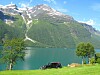 Telt og bil ved en innsjø med høye fjell i bakgrunnen på en solrik dag.