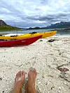 Rød og gul kajakk på en strand med utsikt mot fjell og stille hav.