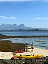 Person på strand med gule kajakker i forgrunnen og fjell i bakgrunnen.