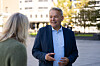 Mann i blå dress snakker med en kvinne utendørs foran en bygning.