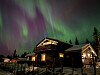 Nordlys i grønt og lilla over en opplyst hytte ved Sjusjøen om kvelden.