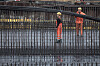 To arbeidere i verneklær og hjelmer står bak armeringsjern på en byggeplass.