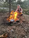 Person sitter ved et bål i skogen med gryte og kokeplate blant steiner og barnåler.