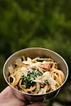 Hånd som holder en metallskål med pasta, urter og revet ost.