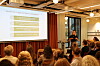 Foredragsholder ved skjerm presenterer for et sittende publikum i et konferanserom.