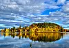 Skogkledd øy med gule høsttrær speiler seg i en innsjø under blå og skyet himmel.